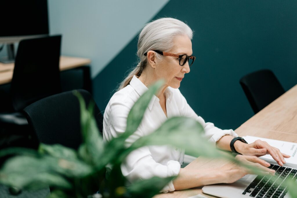 pexels-photo-5971257-5971257 Confident senior woman with glasses working on a laptop in a modern office setting.